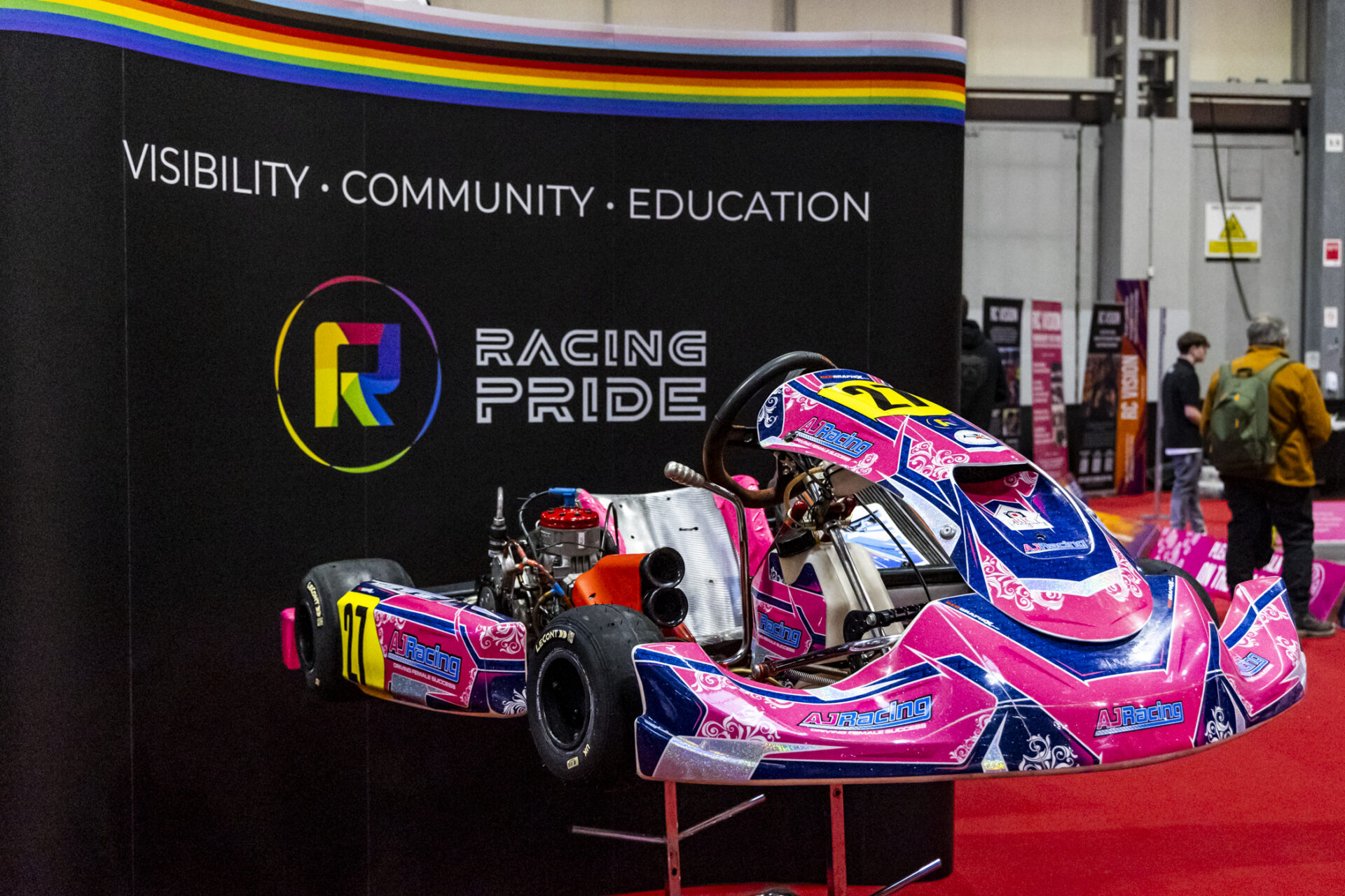 Pink and blue kart from AJ Racing team is shown on display in front of black signage with the Racing Pride logo.