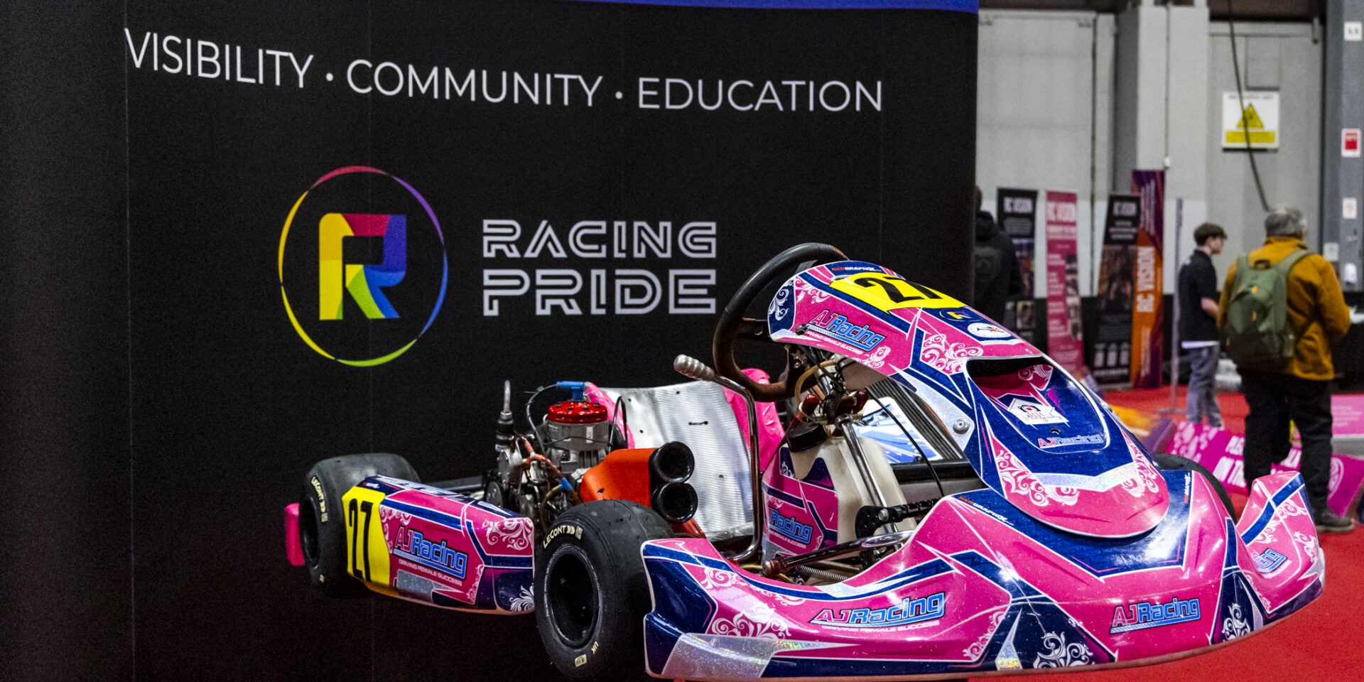 Pink and blue kart from AJ Racing team is shown on display in front of black signage with the Racing Pride logo.