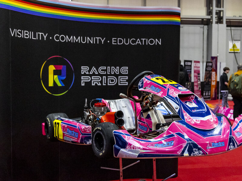 Pink and blue kart from AJ Racing team is shown on display in front of black signage with the Racing Pride logo.