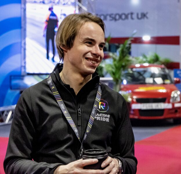 A smiling person (CEO Richard Morris) wearing a black shirt with Racing Pride logo is stood in front of a red car display. Motorsport UK signage is visible in the background.