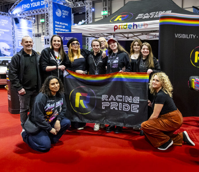 A group of smiling people stand and kneel holding a black flag with rainbow stripes and Racing Pride logo.