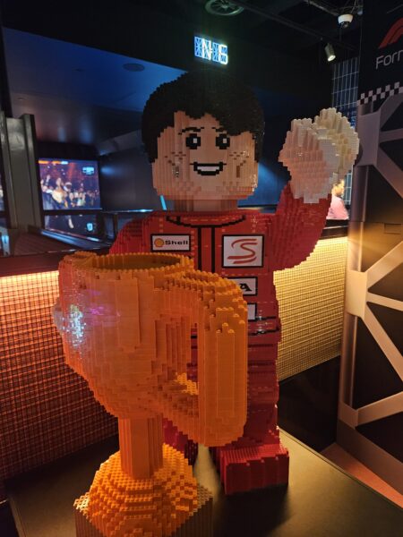 A large male race driver figure with black hair and a red race suit is made out of lego bricks, holding a trophy also made from lego.