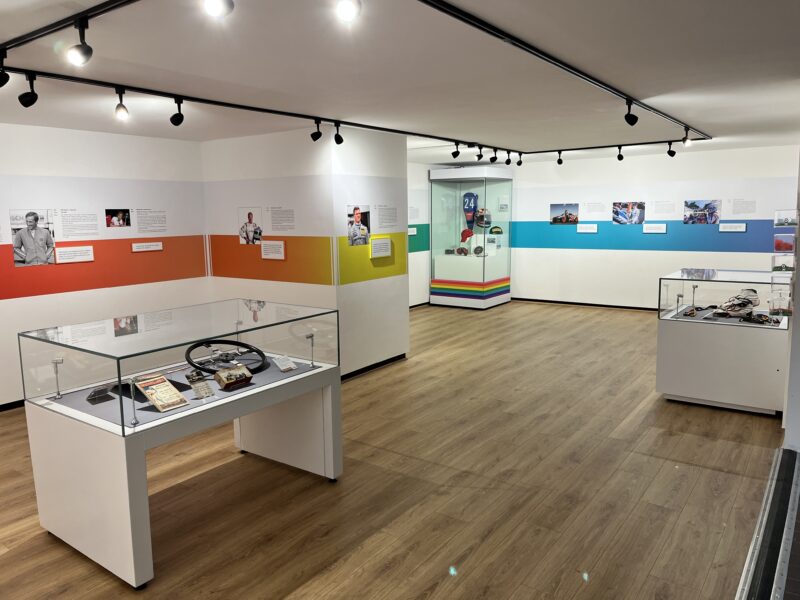 A room with white walls, a series of display cases with glass covers and a rainbow line running along the wall. This highlights pictures of notable LGBTQ+ figures in motorsport history, with artefacts on display including photographs, race suits, helmets and a steering wheel.