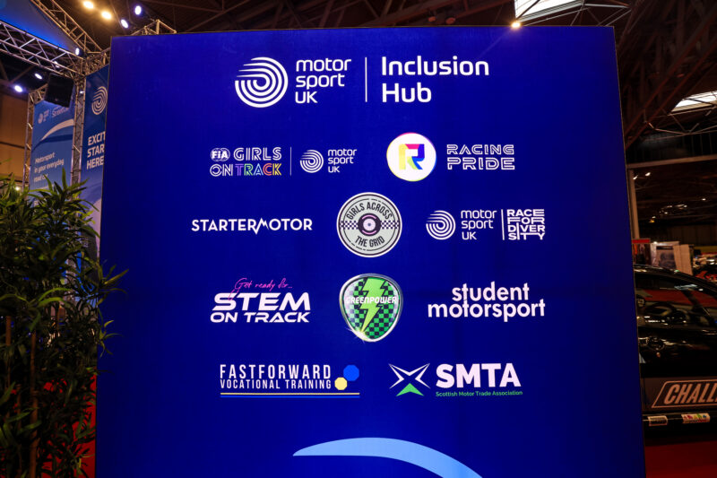 A large blue display sign features the logos and names of several organisations within the Motorsport UK Inclusion hub, including Racing Pride.