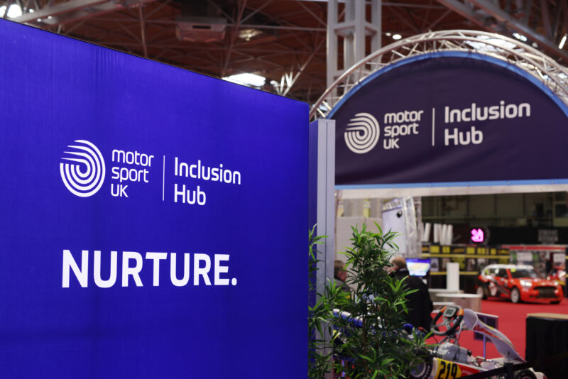 A large blue arched sign with the Motorsport UK log reads 'Inclusion Hub' in the foreground is another sign reading 'Nurture'. The backdrop is the convention centre venue for ASI 2025.